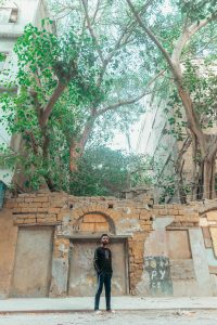 Homme debout contre un mur rustique sous des arbres imposants en milieu urbain, incarnant l'exploration.