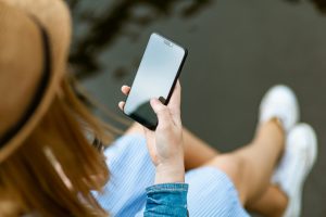 Une jeune femme tenant un smartphone en étant assise en extérieur en été, capturant un moment de détente et d'utilisation de la technologie.