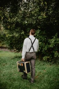 Un homme en bretelles marchant dans une zone herbeuse avec une valise vintage.