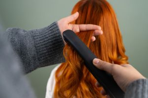 Hands styling long red hair using a flat iron indoors. Haircare and beauty concept. Mains coiffant de longs cheveux roux avec un lisseur à l'intérieur. Concept de soin et de beauté des cheveux.