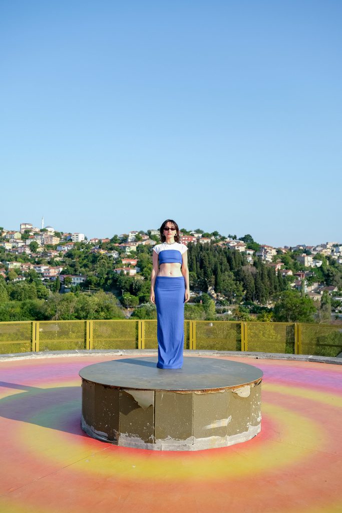 Une femme en tenue bleue pose en extérieur sur une plateforme avec une ville pittoresque en arrière-plan.