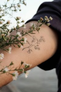 Un gros plan d'un tatouage floral entouré de fleurs blanches sur le bras d'une personne, évoquant une ambiance naturelle et artistique.