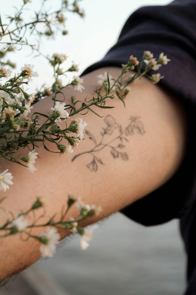 Un gros plan d'un tatouage floral entouré de fleurs blanches sur le bras d'une personne, évoquant une ambiance naturelle et artistique.