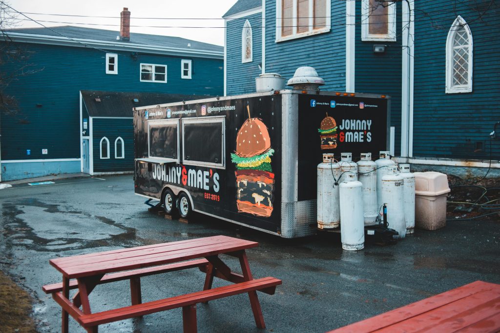 From above of food truck placed on wet asphalt area with wooden tables and benches in front of blue buildings Camionnette de nourriture vue d'en haut, placée sur une zone asphaltée humide avec des tables et bancs en bois devant des bâtiments bleus