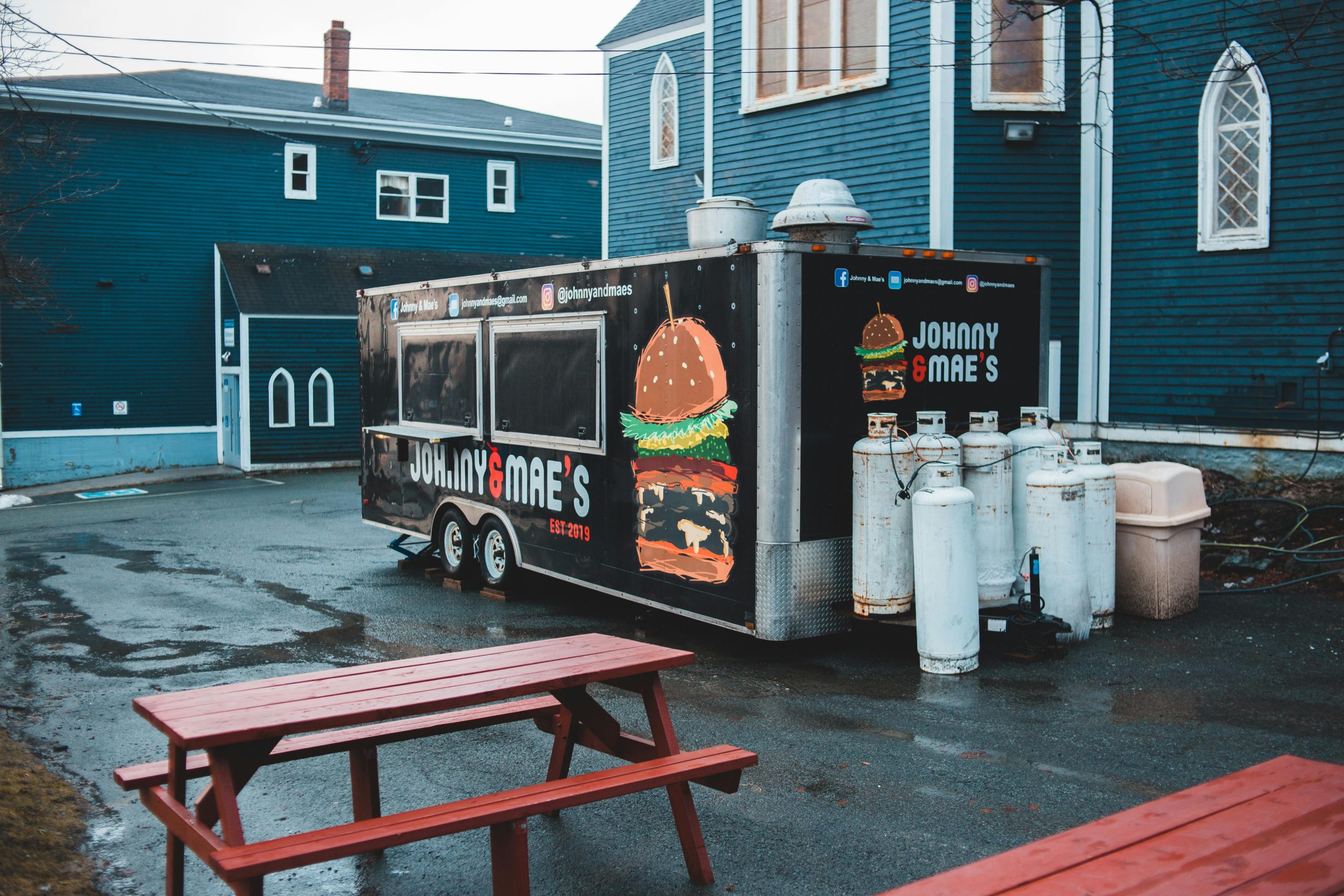 From above of food truck placed on wet asphalt area with wooden tables and benches in front of blue buildings Camionnette de nourriture vue d'en haut, placée sur une zone asphaltée humide avec des tables et bancs en bois devant des bâtiments bleus