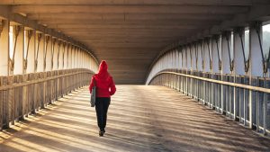 Une femme en manteau rouge traverse un pont moderne, avec la lumière du soleil projetant de longues ombres.