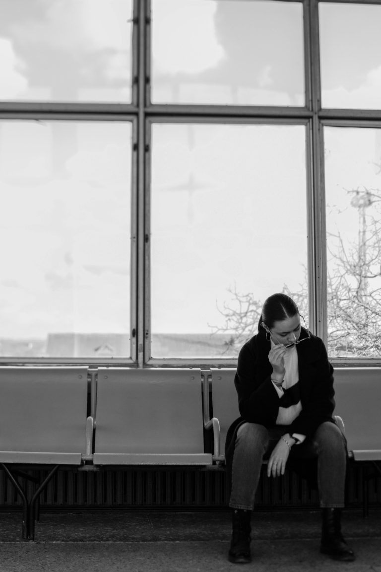 Black and white image of a woman wearing sunglasses, seated in a waiting area looking contemplative. Image en noir et blanc d'une femme portant des lunettes de soleil, assise dans une salle d'attente et regardant de manière contemplative.