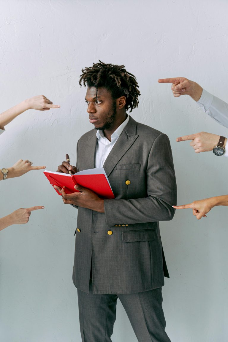Businessman holding a red notebook surrounded by pointing hands. Concept of pressure and decision-making. Homme d'affaires tenant un carnet rouge entouré de mains pointées. Concept de pression et de prise de décision.