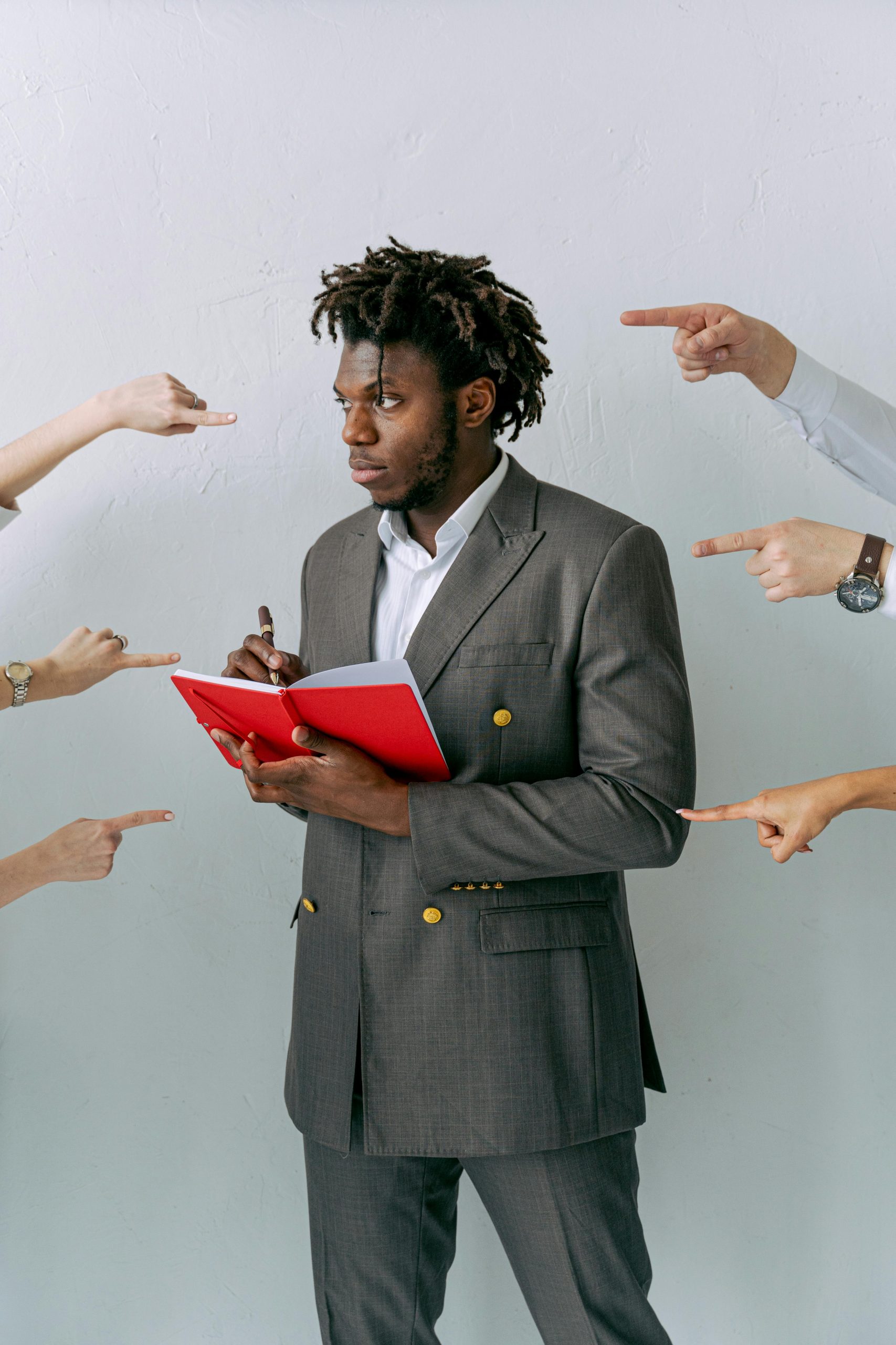 Businessman holding a red notebook surrounded by pointing hands. Concept of pressure and decision-making. Homme d'affaires tenant un carnet rouge entouré de mains pointées. Concept de pression et de prise de décision.