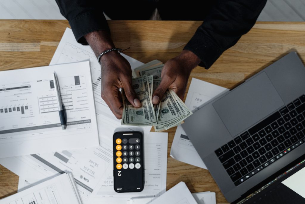 Person counting US dollars, using a calculator and laptop, with financial documents on a wooden desk. Une personne compte des dollars américains, utilisant une calculatrice et un ordinateur portable, avec des documents financiers sur un bureau en bois.