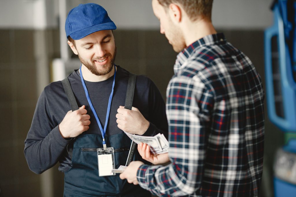 A mechanic in a blue cap receives cash payment from a customer in a plaid shirt, smiling. Un mécanicien en casquette bleue reçoit un paiement en espèces d'un client en chemise à carreaux, souriante.
